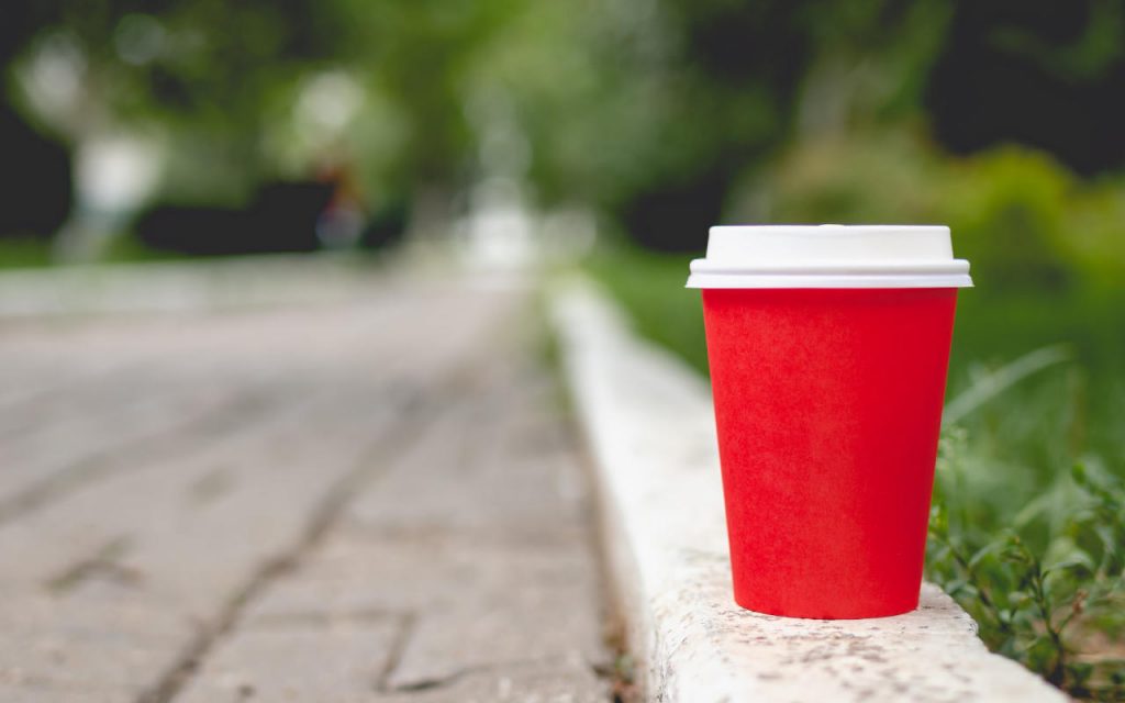 A red disposable coffee cup with white plastic lid on, placed on the kerb in a park.