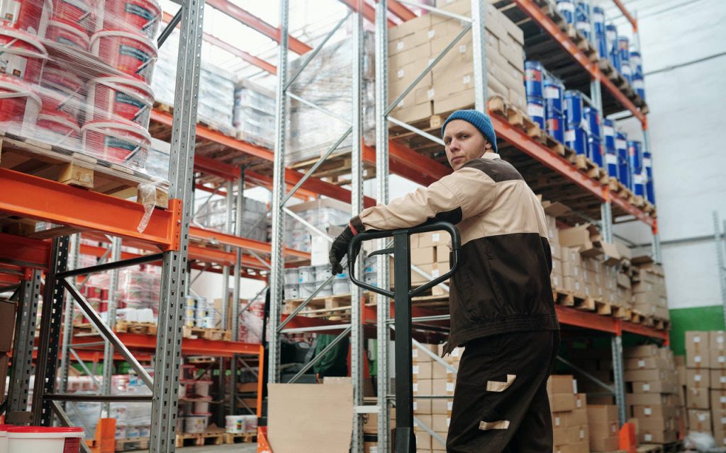 Warehouse worker posing by pallet truck, polythene wrapped goods visible on shelves in background