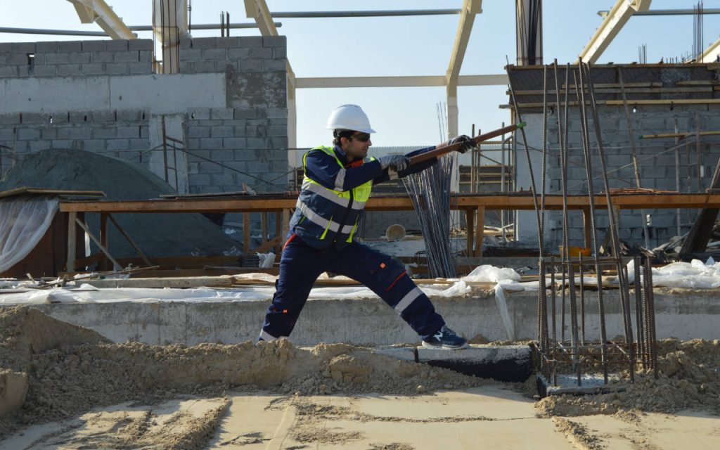 Man working on construction site, pulling on rebar. Polythene sheeting can be seen on the ground, seemingly used as a temporary cover for concrete. The building is in progress and largely incomplete, the image appears to be outdoors in full daylight.