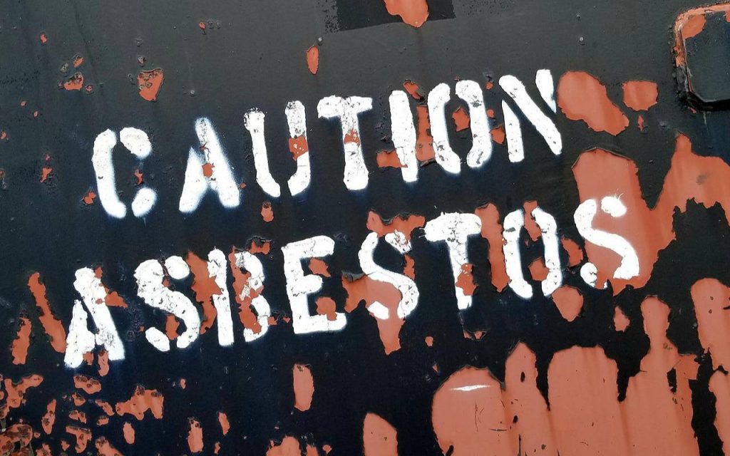 Closeup photograph of a sign that reads "CAUTION ASBESTOS" in capital letters, printed with white paint and stencil onto a rusting piece of metal. Orange and black paint is flaking.