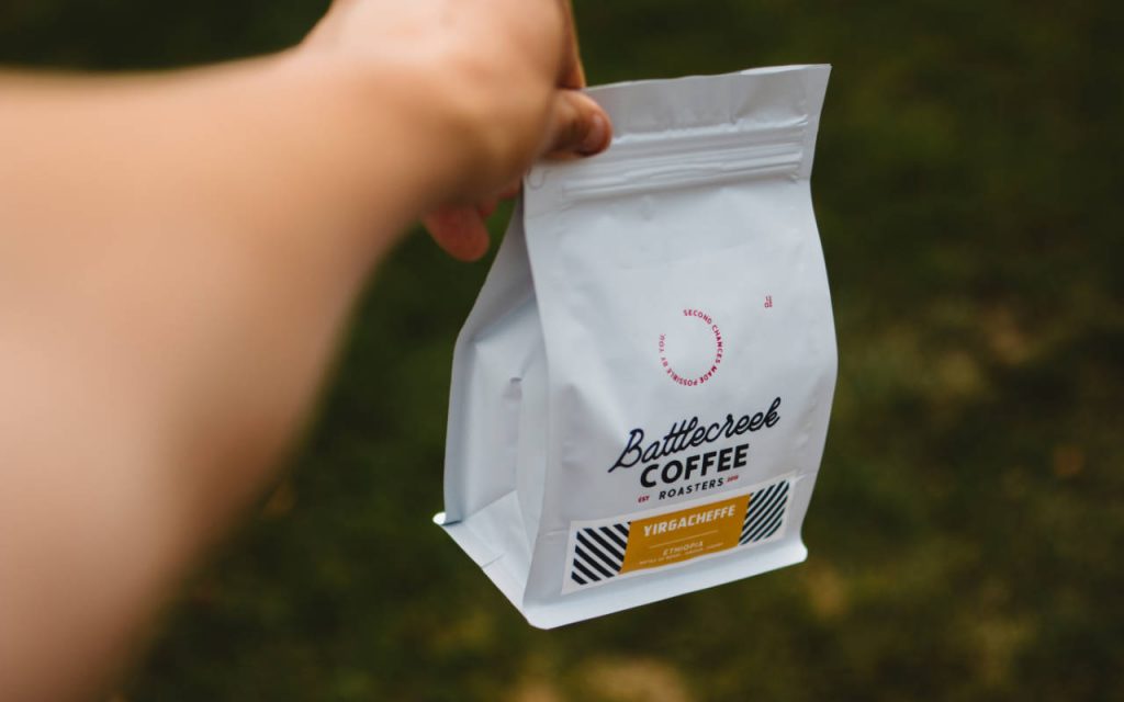 Shallow focus photography of a bag of coffee, held above grass by a left hand at arm's length. The bag is white, labelled with black lettering reading "Battlecreek Coffee", and there is an orange box with more information below. The bag is bottom gusseted, with a click seal top.