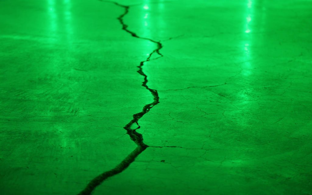 A professional photograph of concrete flooring with a large crack running through it. Focus is soft in the background, and thew whole image is bathed in green light. The texture of the concrete is visibly smooth, save for the large, wide crack that occupies the left half of the image.