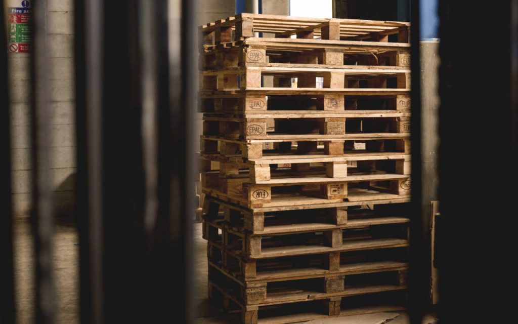 Pallets in a warehouse, stacked up high. Daylight casts moody shadows on the scene.