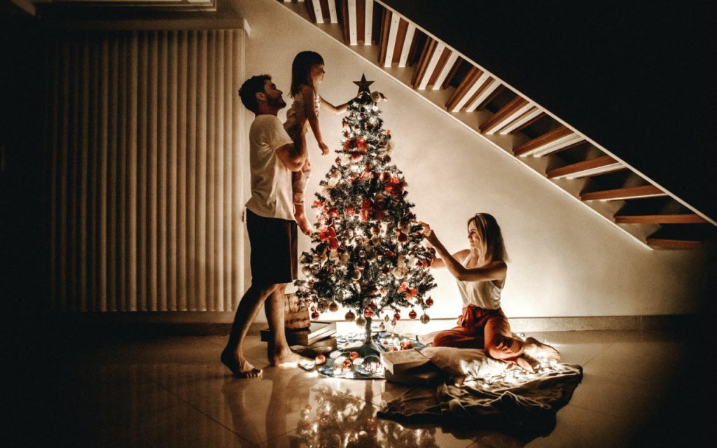 A family of three decorating an artificial Christmas tree. They are under the stairway in their home, illuminated only by the fairy lights on the tree. A father lifts his child up to place the star on top. A mother arranges decorations.