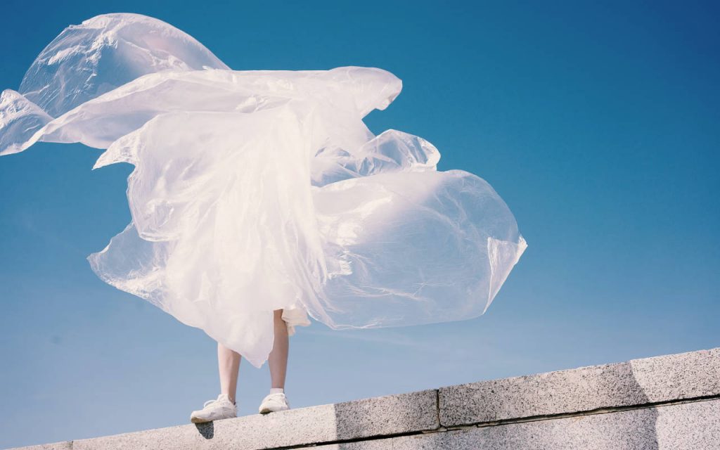 Surreal photograph of a person holding polythene sheeting, while stood on a rooftop, against a blue sky. The person is fully shrouded in polythene film, with only their shins and feet visible. The bright sunlight shining on the subject is dispersed by the translucent sheeting, making it appear white, and making it look like the person has disappeared.