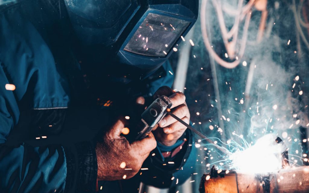 Close up shot of an engineer welding, while wearing protective gear. Hands are exposed and visible. The sparks suggest arc welding, but could be another weld.