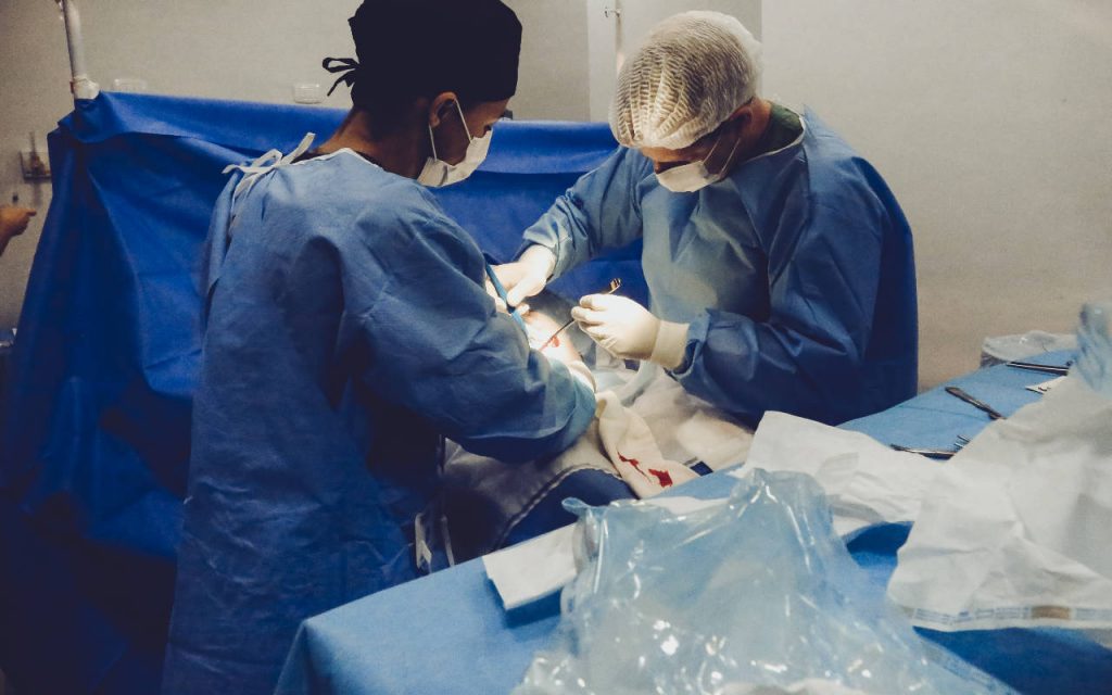 A surgeon and assistant use medical plastics during surgery. They are wearing blue scrubs. A medical polythene bag can be seen in the foreground. A blue sheet is suspended over the patient. The procedure is not shown.