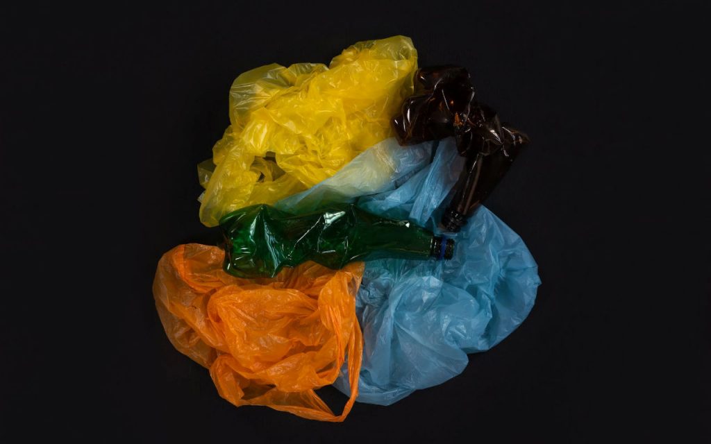 An image of recycled polythene products o na black background. Coloured polythene bags in yellow, blue and orange – plus two plastic bottles in brown and green – all screwed up together.