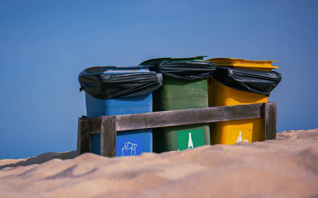 Zoomed photo of black polythene bags lining coloured bins on a sandy beach. The sky is vivid blue, and sand can be seen in foreground. The bins and polythene bags are in sharp focus. They are in a wooden frame.