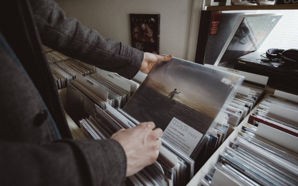 Inside a record shop, a man in a tweed jacket holds a vinyl record in a polythene sleeve. We see the image from a third person's perspective, almost over the man's shoulder. His head and face are not visible. The record is in full view – Pink Floyd, The Endless River. The cover shows a small boat being paddled in a vast body of water, into a sunset with dull hues and dramatic clouds.