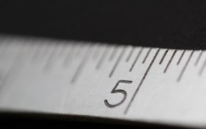Closeup image of a tape measure at 5 inches. the tape is white, and the text is black – the texture is visible. The background is black and very out of focus.