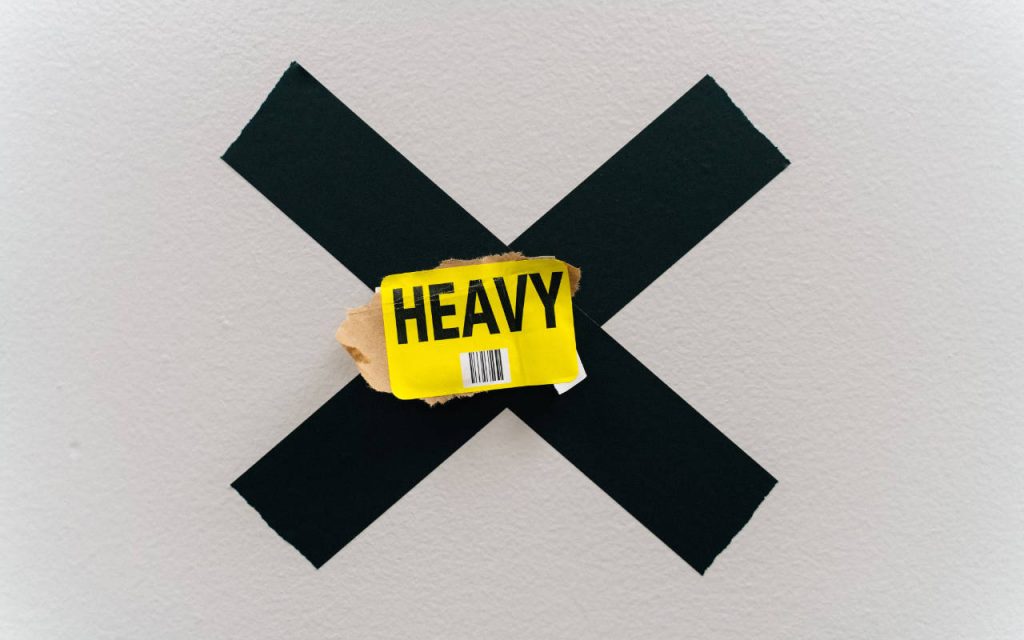 Image of a yellow sticker with thick black txt that reads HEAVY, stuck to a cross of black duct tape, stuck to a white, textured wall