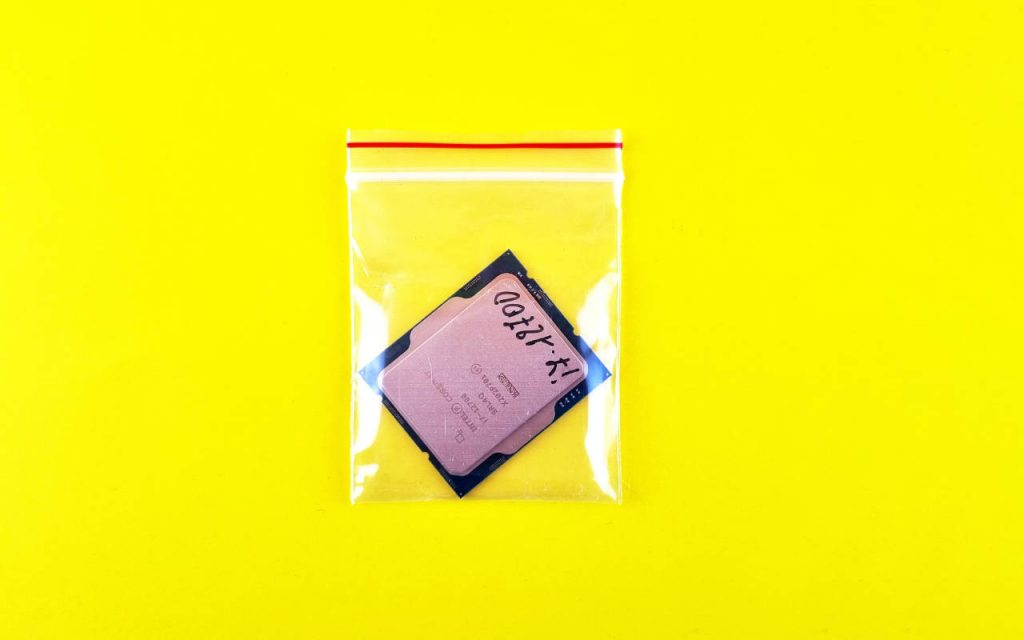 A grip seal polythene bag with a CPU inside, on a vivid, highlighter yellow background. The image is intensely bright. The CPU in the grip seal bag is an Intel Core i7, marked with black Sharpie. It sits nearly upside down in the grip seal bag, at a jaunty angle. There is a red stripe above the grip seal, which is very stark against the vivid yellow backing.