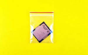 A grip seal polythene bag with a CPU inside, on a vivid, highlighter yellow background. The image is intensely bright. The CPU in the grip seal bag is an Intel Core i7, marked with black Sharpie. It sits nearly upside down in the grip seal bag, at a jaunty angle. There is a red stripe above the grip seal, which is very stark against the vivid yellow backing.