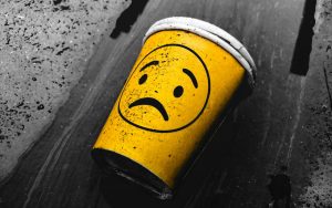 Yellow coffee cups with sad smiley. Shot on a beach, but the background is black and white – only the cup is colour. It's tatty and dirty, but vivid yellow.
