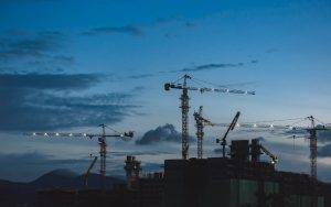 Construction site skyline, seen from a distance. Cranes can be seen against a twilight sky, with a smattering of clouds. Greyish blue tones dominate the moody sky