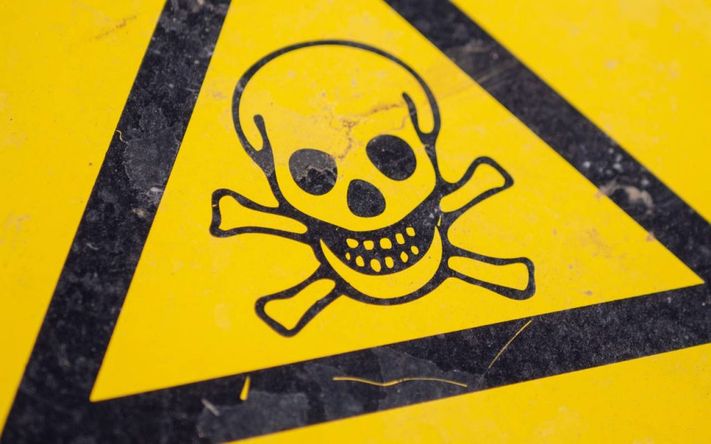 Close up photography of a warning symbol – black skull in a black triangle on yellow background. A close up photograph of signage. Natural patina present, with marks and scratches.